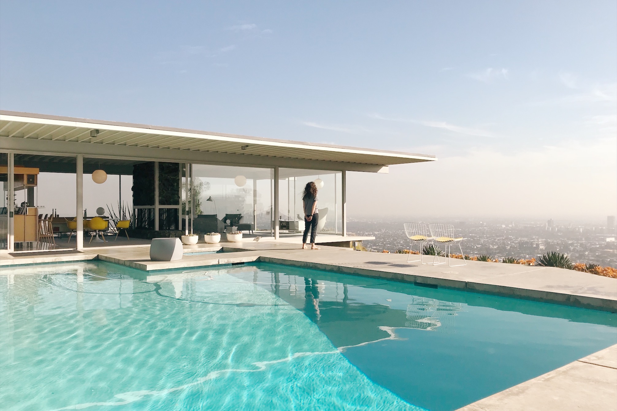 Jessica is standing on the deck of the pool of the iconic Stahl House, overlooking the city of Los Angeles.